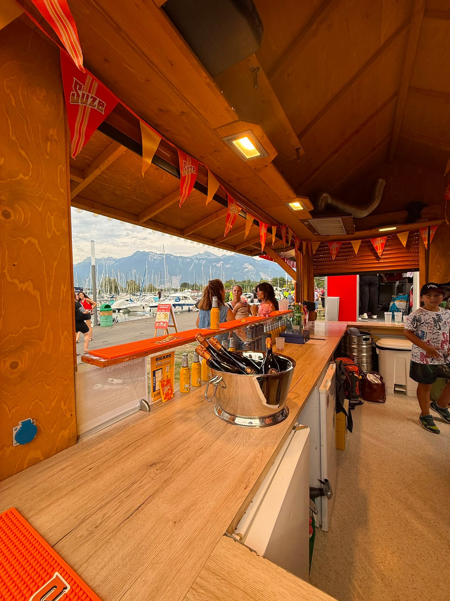 Le bar Suze illuminé en soirée, visiteurs devant le comptoir avant les feux d'artifice
